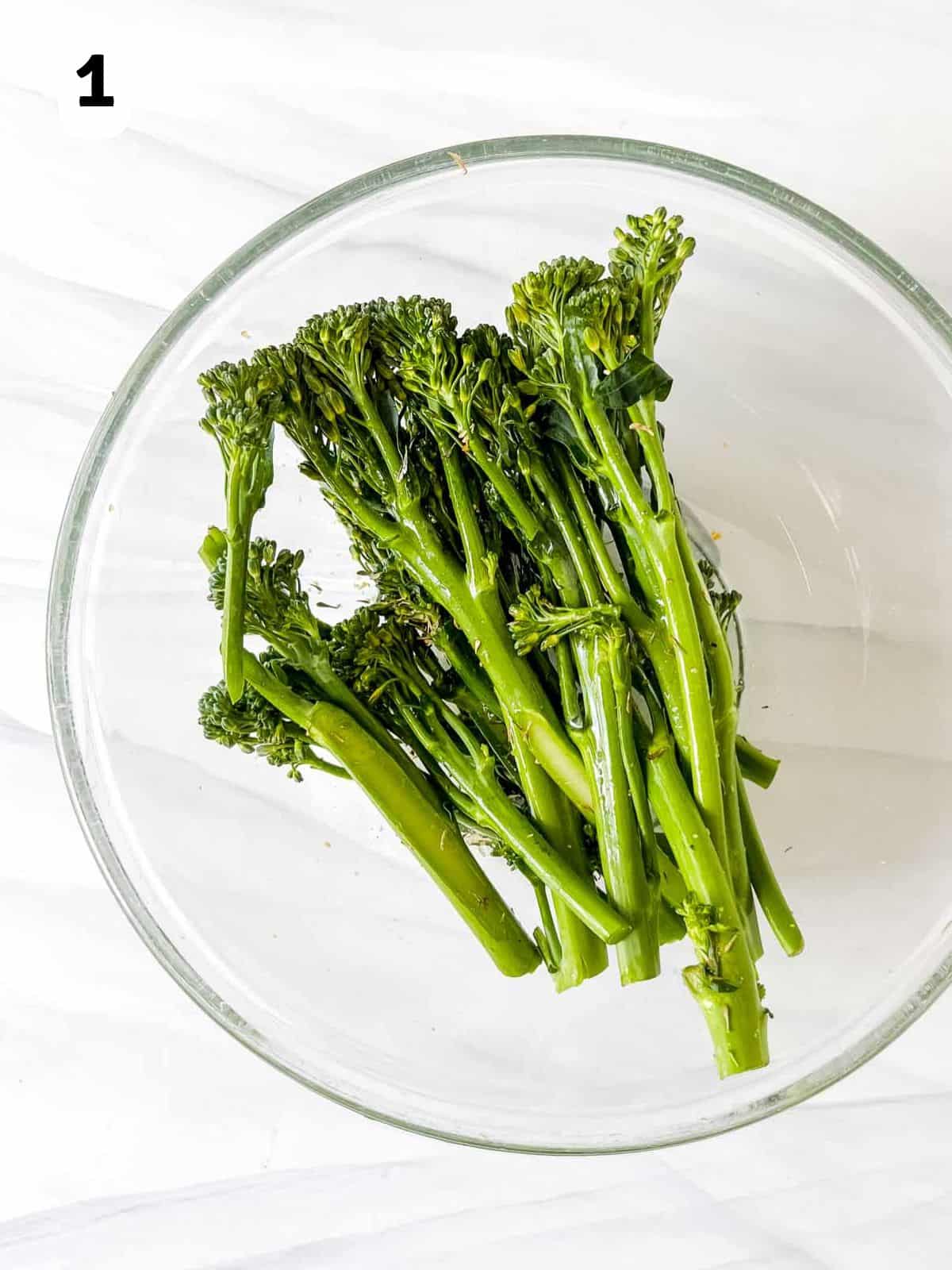 Tenderstem broccoli tossed in olive oil, garlic powder and dried thyme in a glass bowl labelled number one.