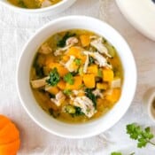 chicken soup with butternut squash in a white bowl next to a pumpkin and fresh parsley.
