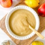 pear applesauce in a white bowl with a wooden spoon next to red apples and pears.