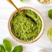 pesto without pine nuts in a light brown bowl with a spoon in it next to fresh basil leaves, olive oil and macadamia nuts.