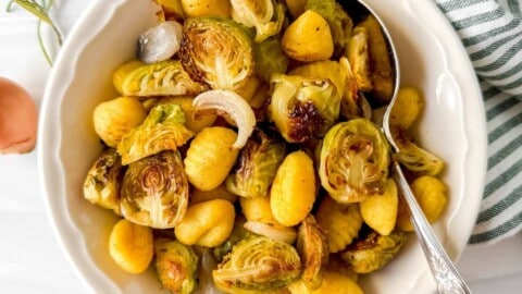 roasted gnocchi and Brussels sprouts in a white bowl with a spoon next to a green striped cloth and shallots.