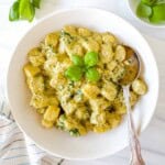 creamy pesto gnocchi in a white bowl with a spoon next to fresh basil leaves.