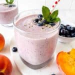 blueberry peach smoothie in a glass next to fresh peaches and blueberries.