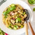 chicken pesto pasta salad in a white bowl next to fresh basil leaves, radishes and salad servers.