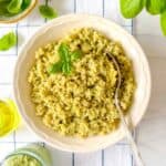 pesto quinoa in a bowl next to a jar of pesto, fresh basil leaves and olive oil.