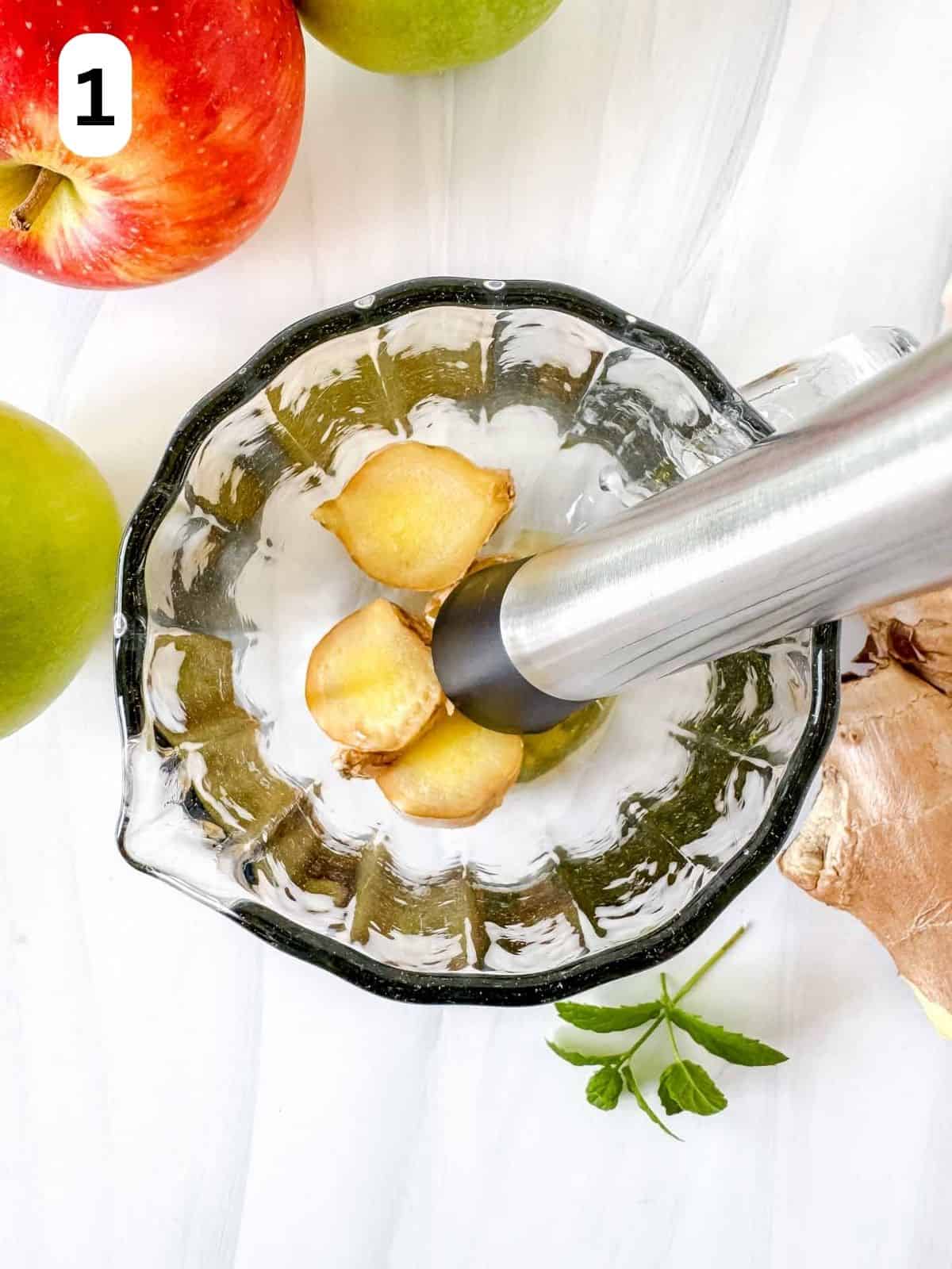 root ginger slices being muddled in a glass jug next to fresh apples labelled number one.