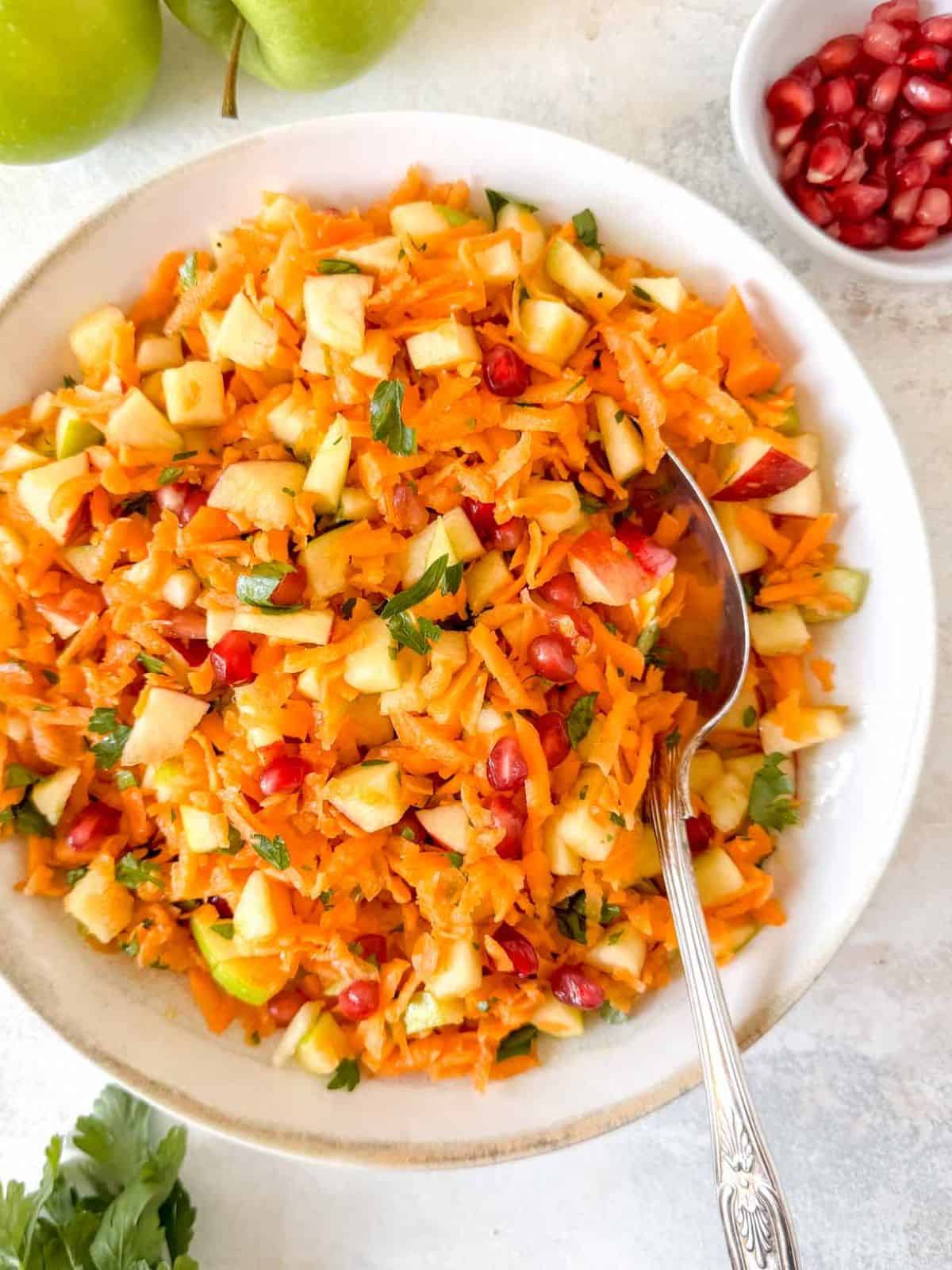carrot and apple salad with pomegranate and parsley in a bowl with a serving spoon.