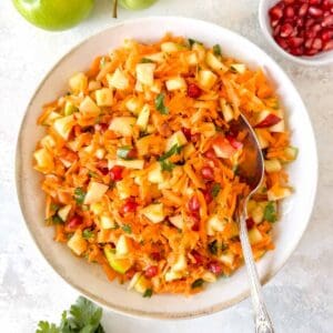carrot and apple salad in a bowl with a serving spoon next to apples and a small bowl of pomegranate arils.