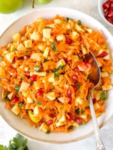 carrot and apple salad in a bowl with a serving spoon next to apples and a small bowl of pomegranate arils.