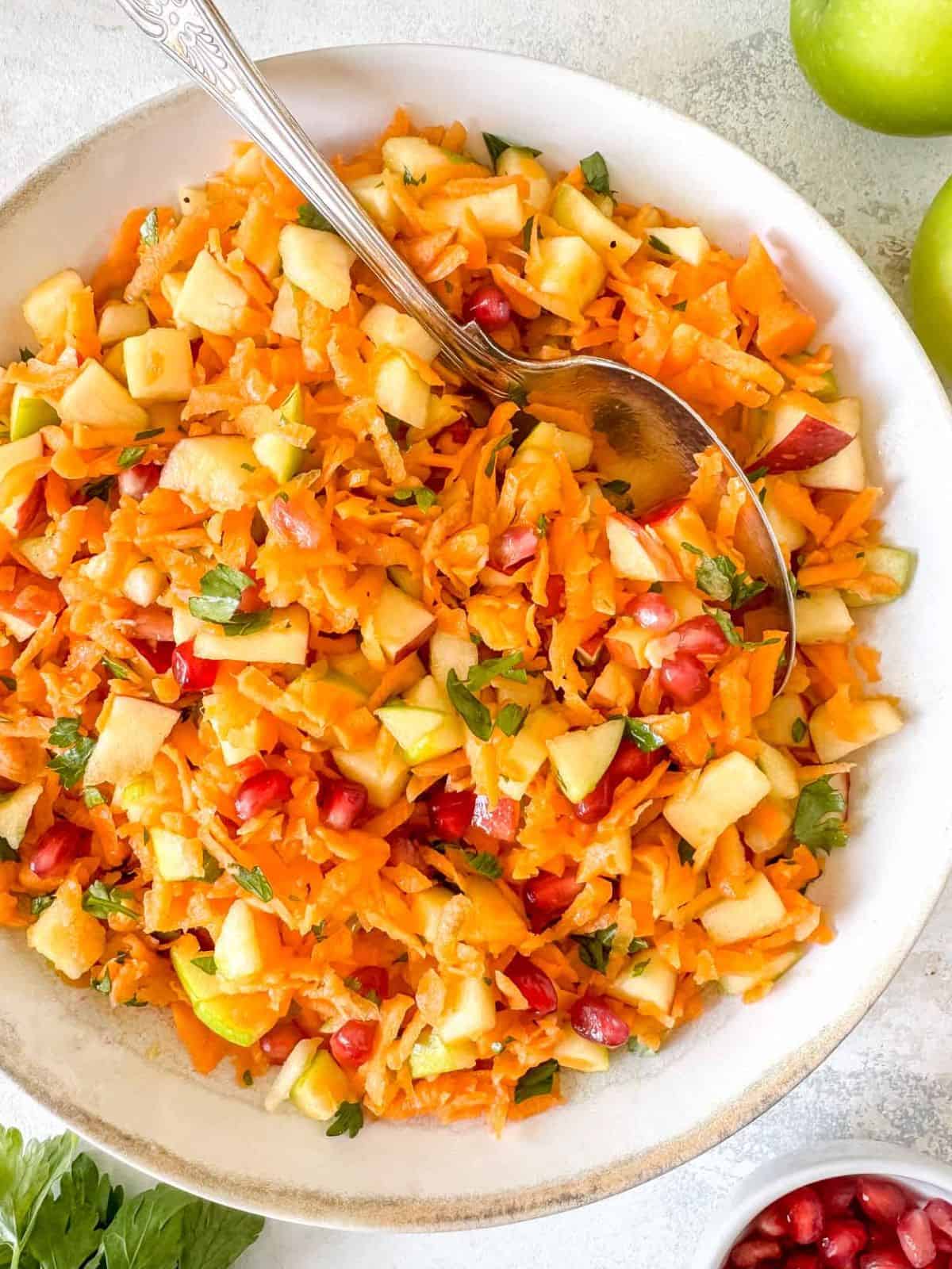 carrot apple salad with pomegranate and parsley in a bowl with a serving spoon.