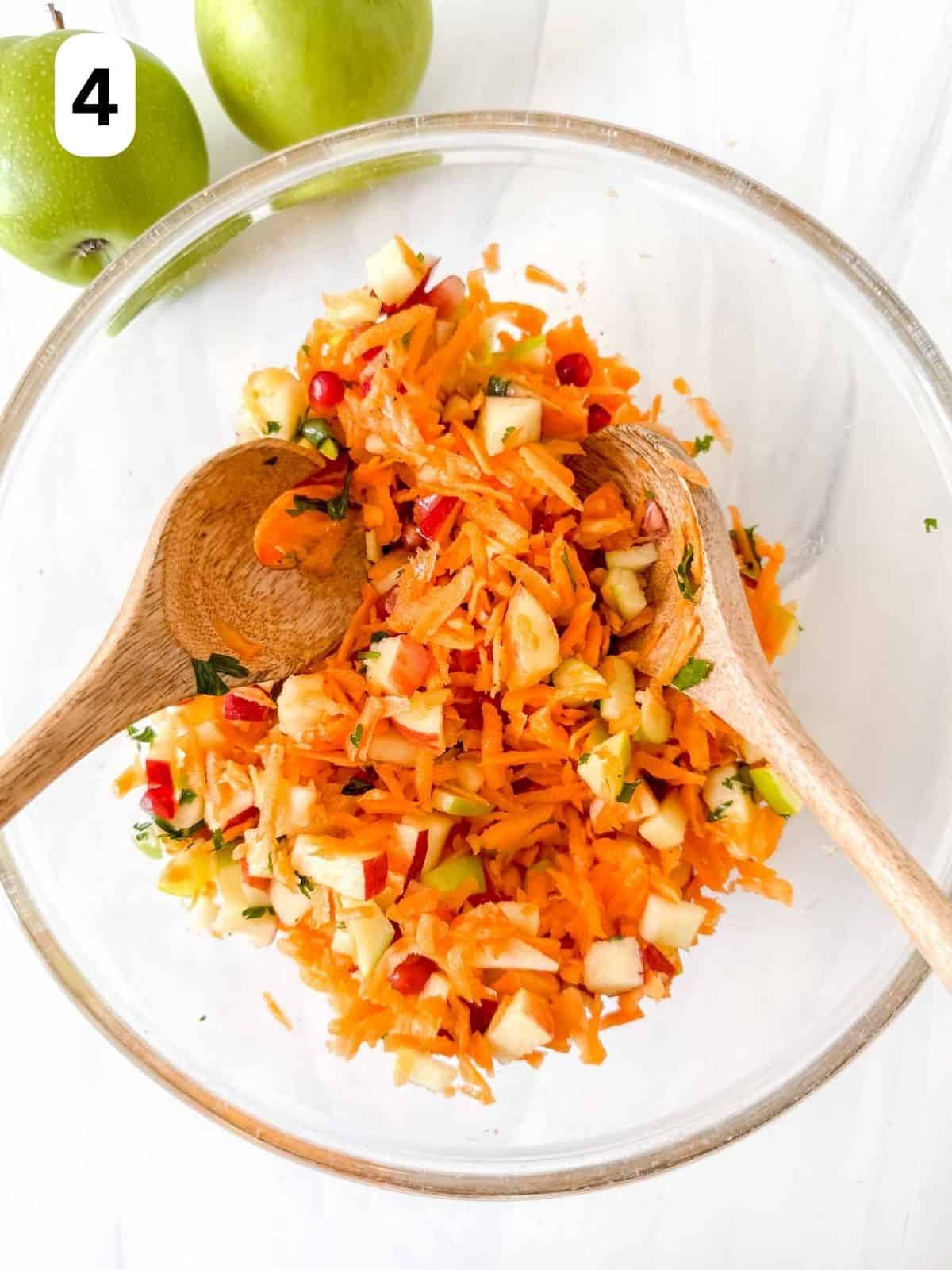 carrot apple salad in a glass bowl being tossed with salad servers labelled number four.