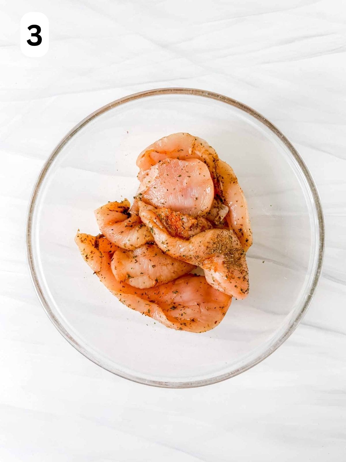 thin sliced chicken breasts coated in olive oil, herbs and spices in a large glass bowl labelled number three.