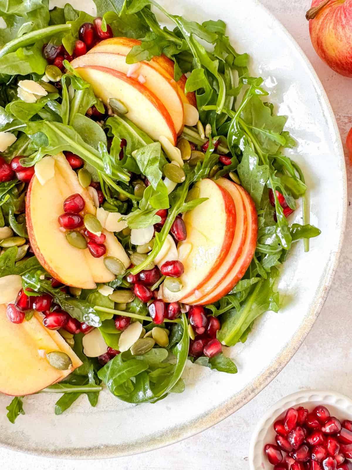 apple and pomegranate salad with arugula in a white bowl next to red apple and a small bowl of pomegranate seeds.