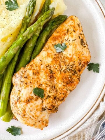 oven baked thin sliced chicken breast on a plate with asparagus and mashed potato garnished with fresh parsley.