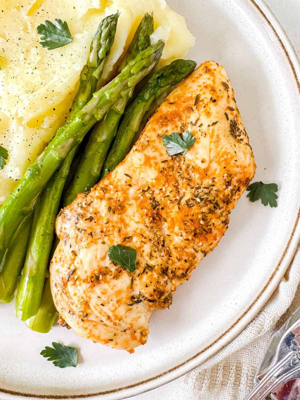 baked thin sliced chicken breast on a plate with mashed potatoes and asparagus.