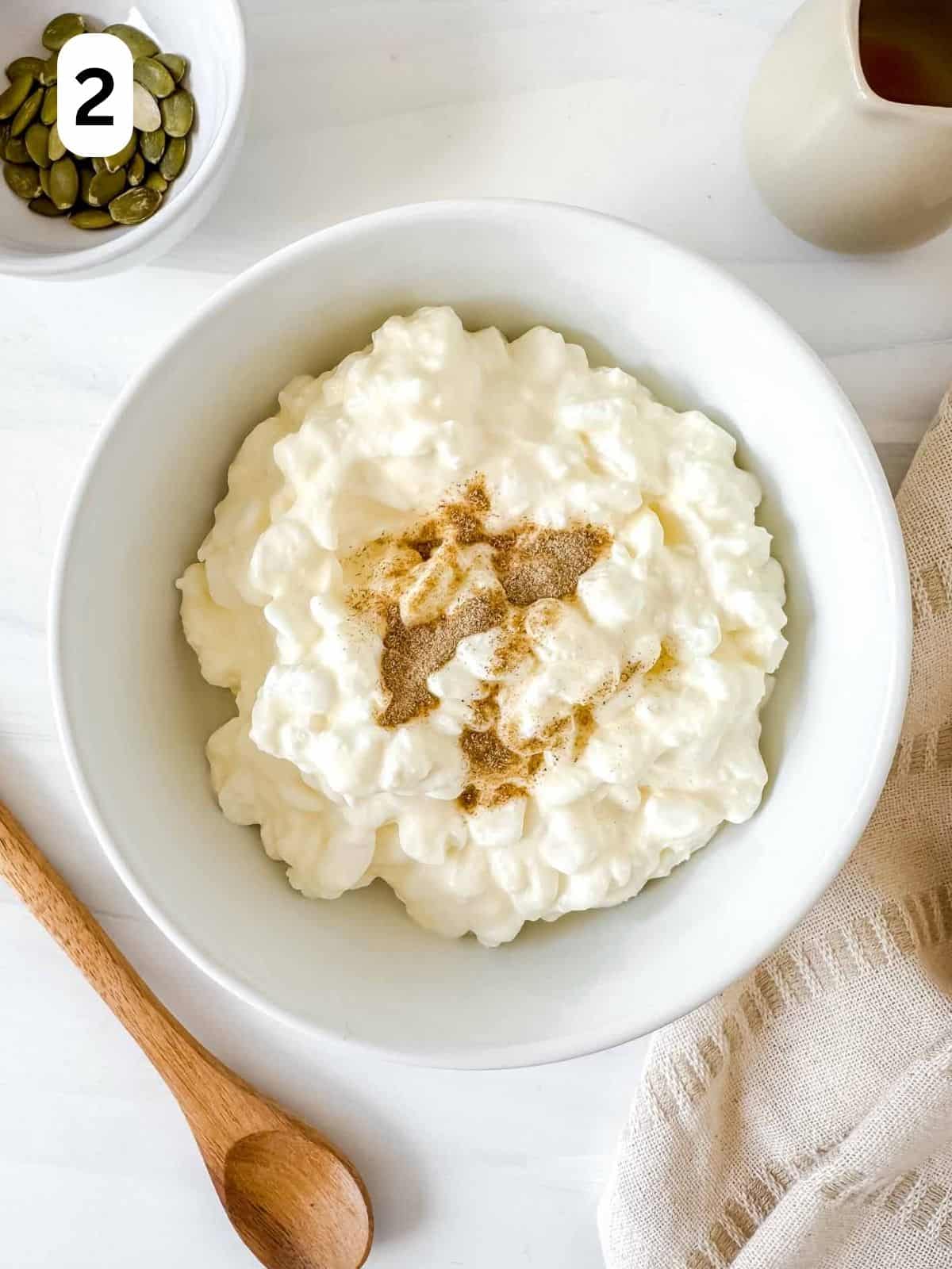 cottage cheese sprinkled with cardamom in a white bowl next to a spoon, cloth and jug.