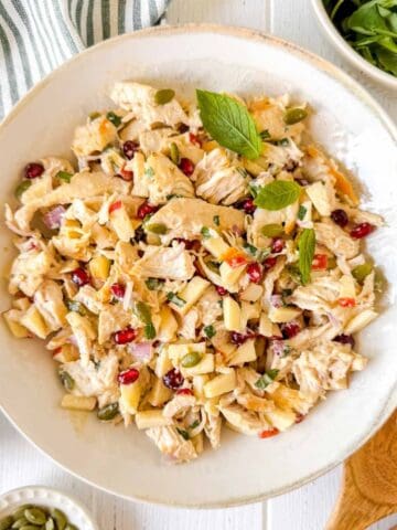 chicken and pomegranate salad in a white bowl next to small bowls of pomegranate seeds and pumpkin seeds.
