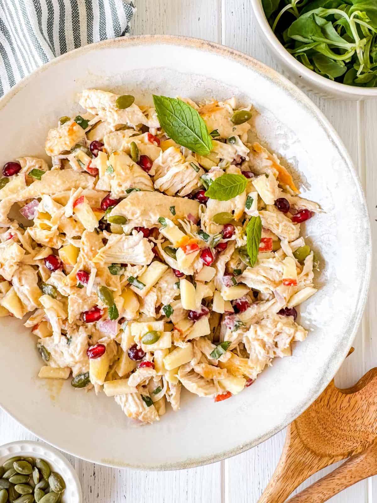 chicken and pomegranate salad with tahini dressing in a white bowl next to salad servers and a bowl of lettuce.