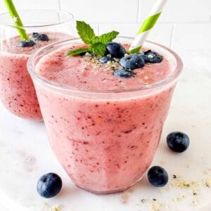 frozen fruit smoothie without yogurt in two glasses topped with blueberries, with a green straw.