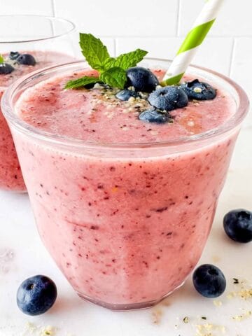 frozen fruit smoothie without yogurt in two glasses topped with blueberries, with a green straw.