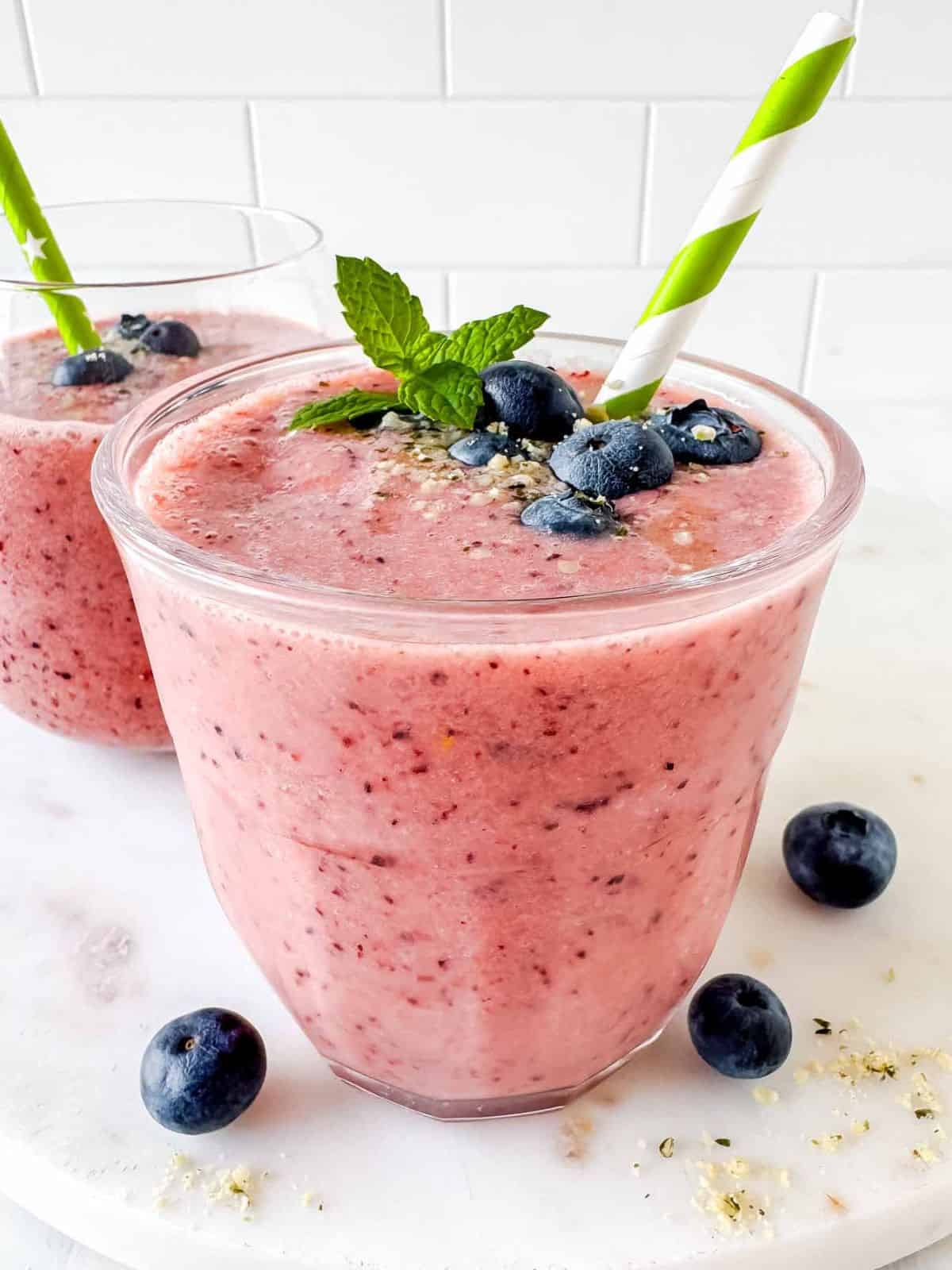 frozen fruit smoothies in two glasses topped with fresh blueberries and a straw.