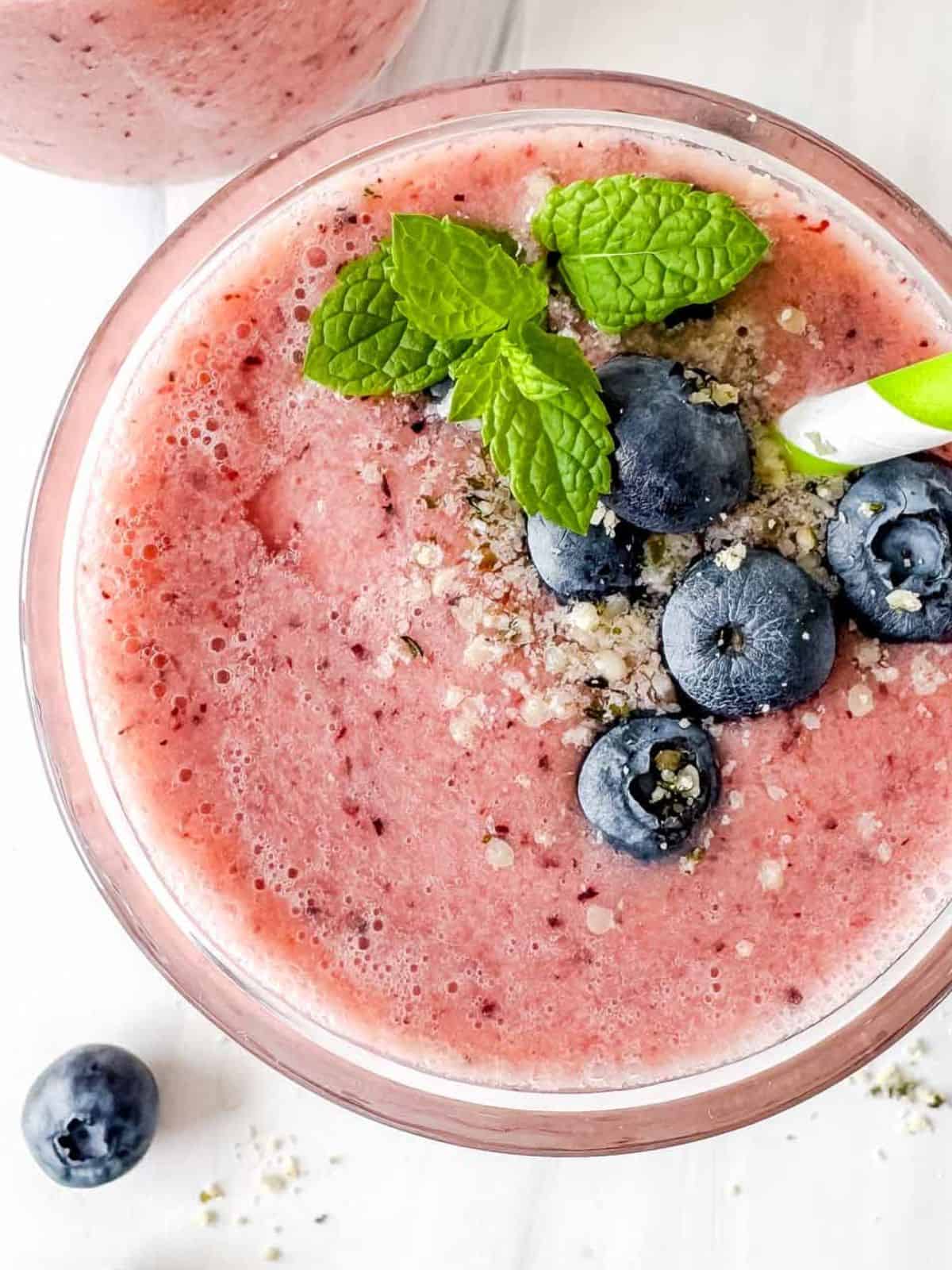 frozen fruit smoothie in a glass topped with blueberries, mint leaves and a straw.