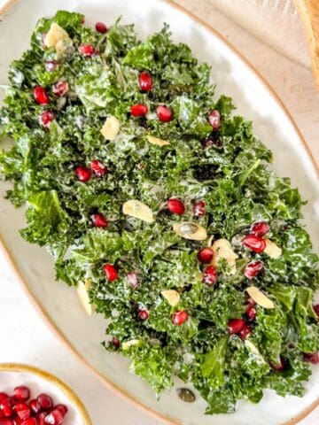 kale and pomegranate salad on a white plate next to a small bowl of pomegranate seeds.