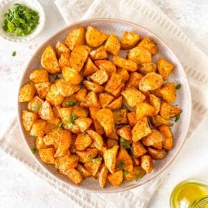 air fryer diced potatoes on a plate next to a small bowl of fresh herbs and a bottle of olive oil.
