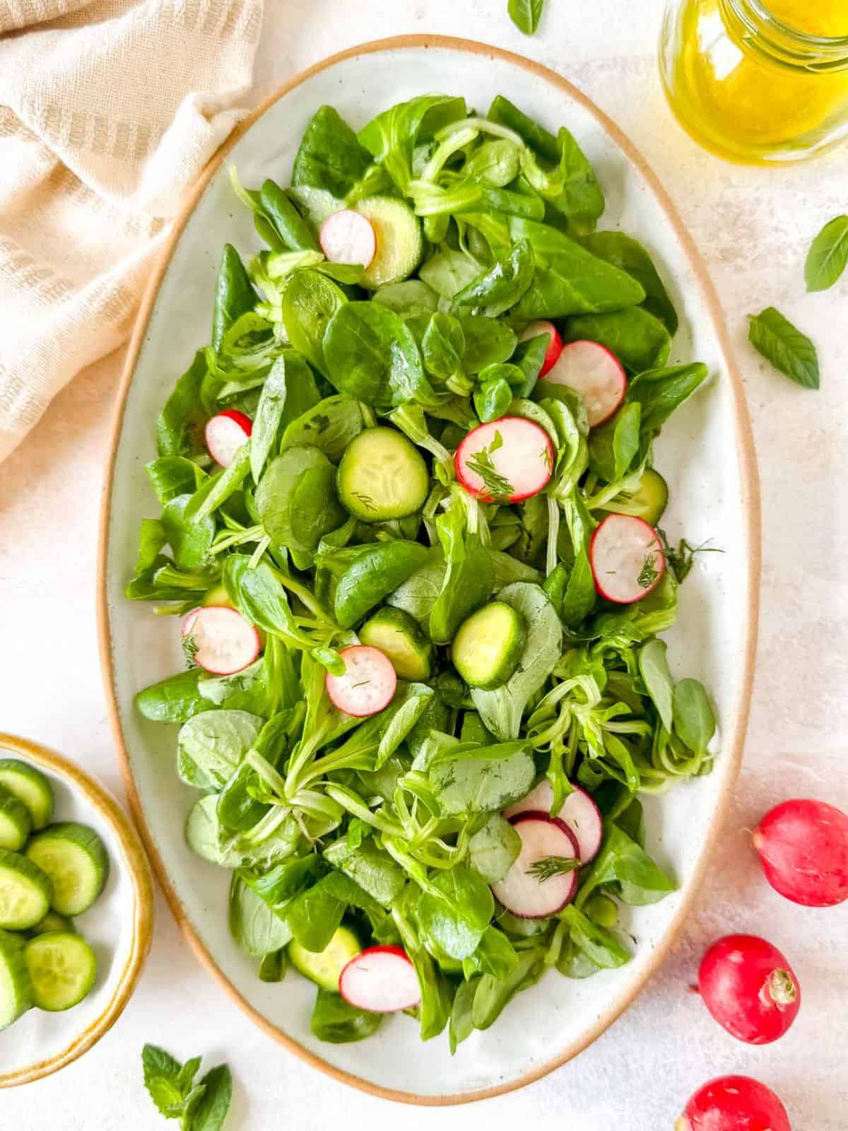 lamb's lettuce salad with cucumbers and radishes on a white plate next to a bottle of olive oil and herbs.