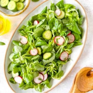 lamb's lettuce salad with mini cucumbers and radishes on a white plate next to salad servers and a bottle of olive oil.