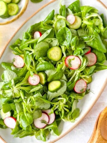 lamb's lettuce salad with mini cucumbers and radishes on a white plate next to salad servers and a bottle of olive oil.