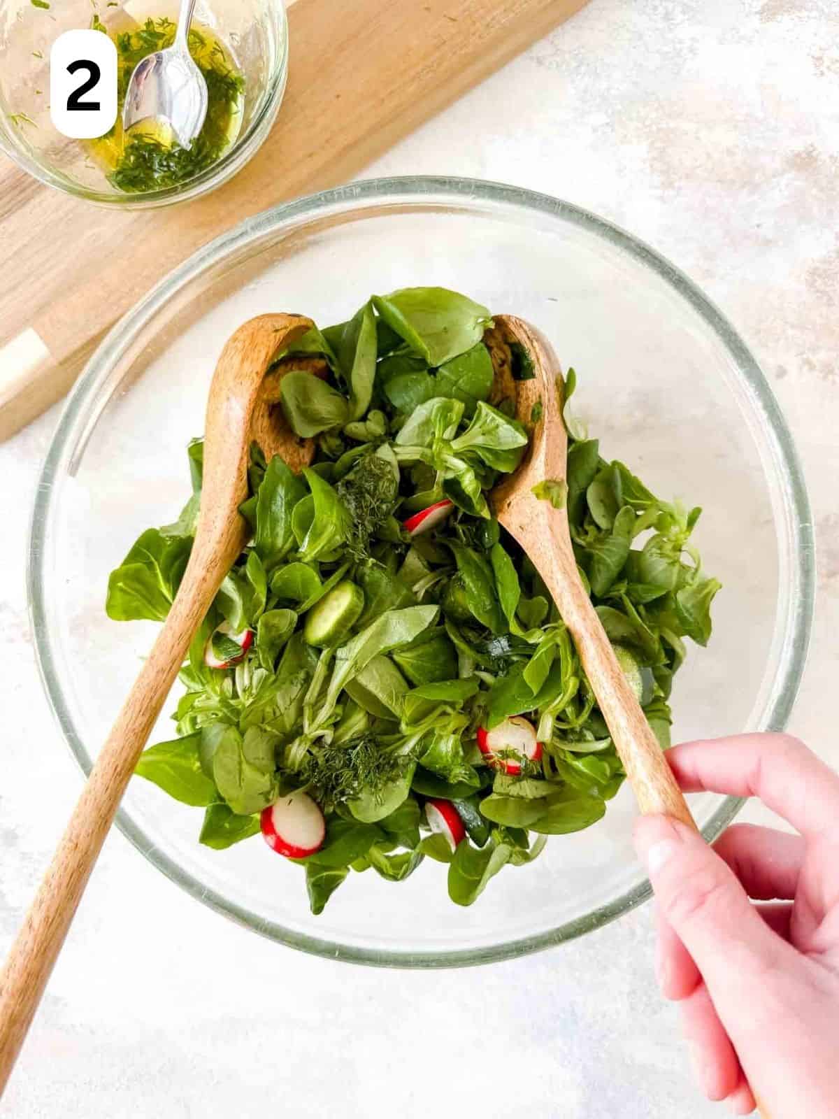 lamb's lettuce, radishes and mini cucumbers in a glass bowl being tossed with salad servers, labelled number two.