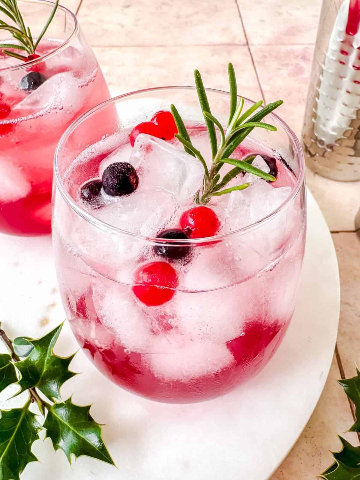 cranberry juice mocktails in two glasses next to a cocktail shaker and rosemary sprigs.