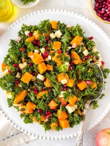 kale butternut squash salad on a white plate next to a small bowl of pomegranate seeds, red apples and olive oil.