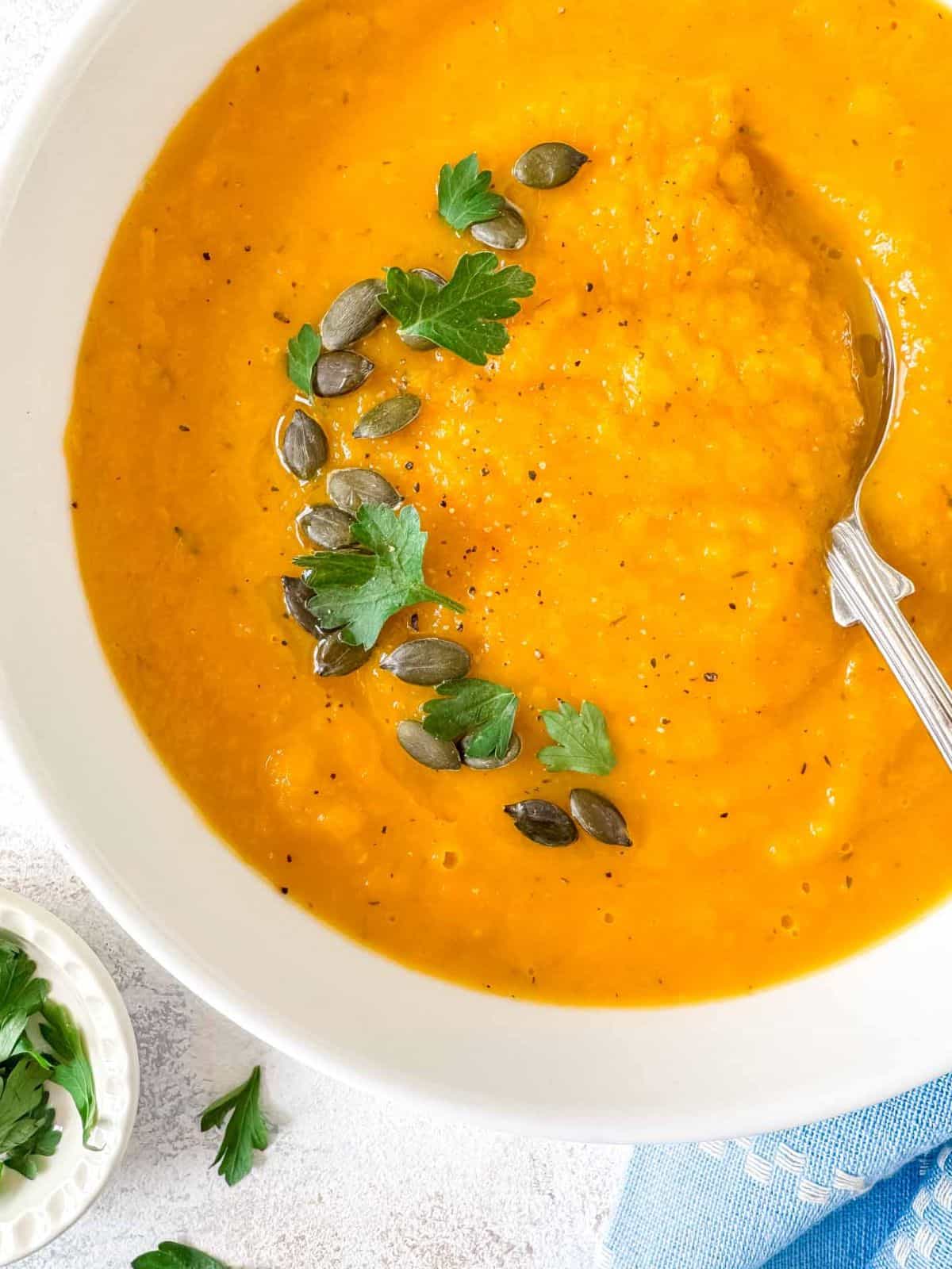 roasted butternut squash and carrot soup in a white bowl garnished with parsley. olive oil and pumpkin seeds next to a small bowl of fresh parsley.