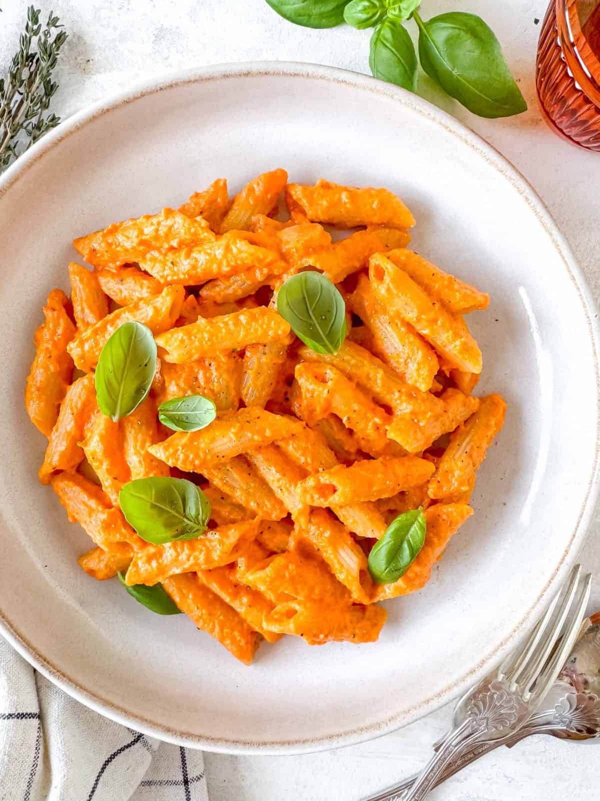 roasted red pepper pasta garnished with fresh basil leaves in a bowl next to an orange glass.