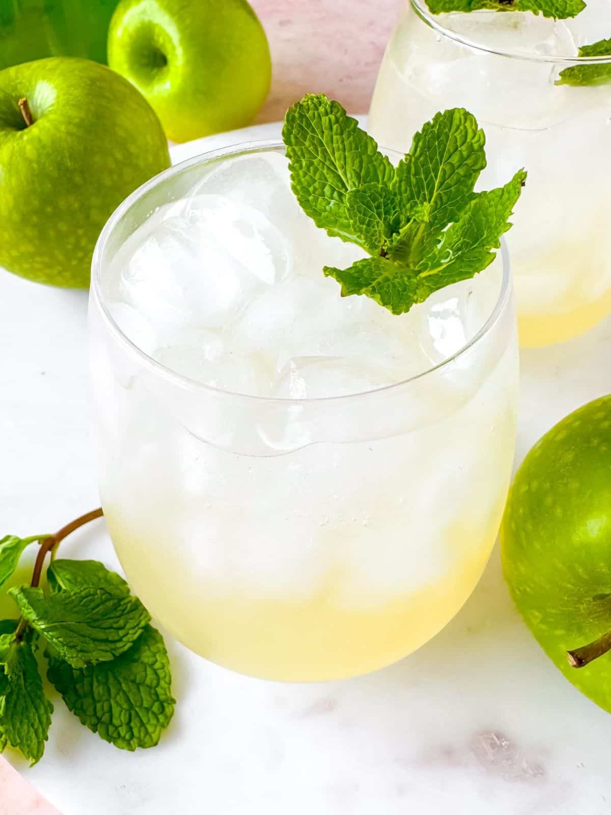 green apple mocktail in two glasses next to Granny Smith apples and mint leaves.