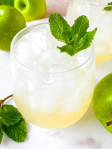 green apple mocktails in two glasses next to Granny Smith apples and mint leaves.