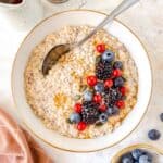 flaxseed overnight oats in a white bowl with a spoon in it topped with fresh berries and maple syrup, next to a small jug and container of ground flaxseed.