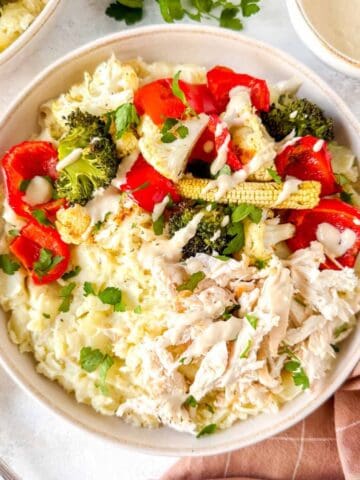 mashed potato bowls with chicken and vegetables drizzled with tahini dressing next to fresh herbs and a brown cloth.