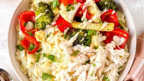 mashed potato bowls with chicken and vegetables drizzled with tahini dressing next to fresh herbs and a brown cloth.