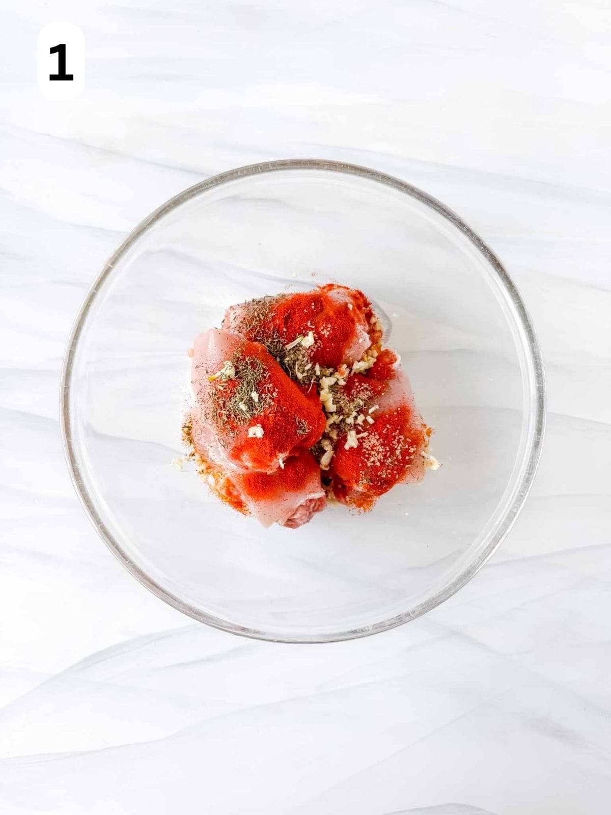 boneless skinless chicken thighs in a glass bowl with olive oil, paprika and herbs sprinkled on top, labelled number one.