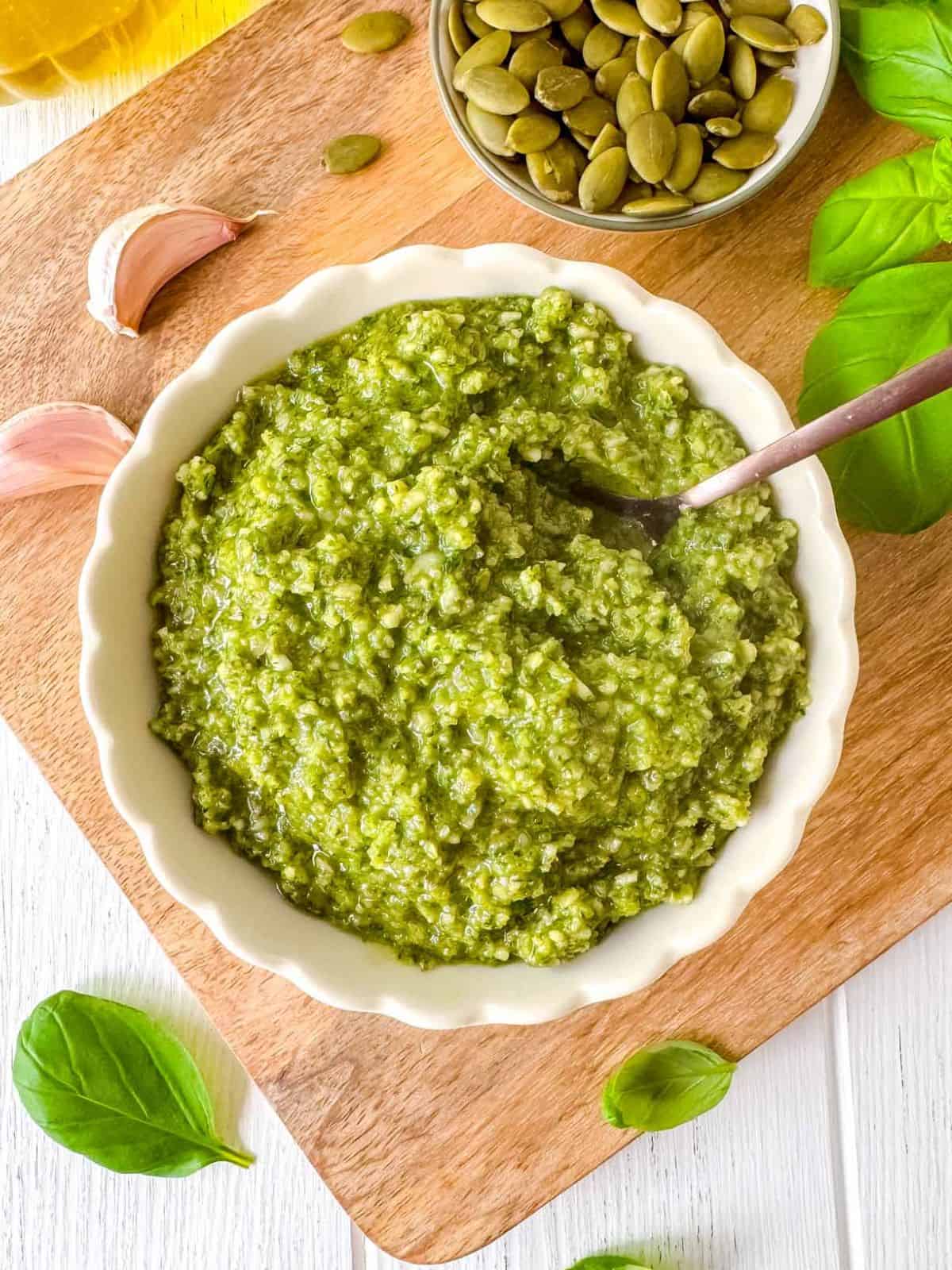 pumpkin seed pesto sauce in a bowl next to pumpkin seeds, fresh basil leaves, garlic and olive oil.