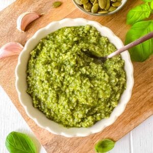 pumpkin seed pesto sauce in a bowl next to fresh basil leaves, garlic and pumpkin seeds.