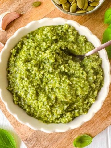 pumpkin seed pesto sauce in a bowl next to fresh basil leaves, garlic and pumpkin seeds.