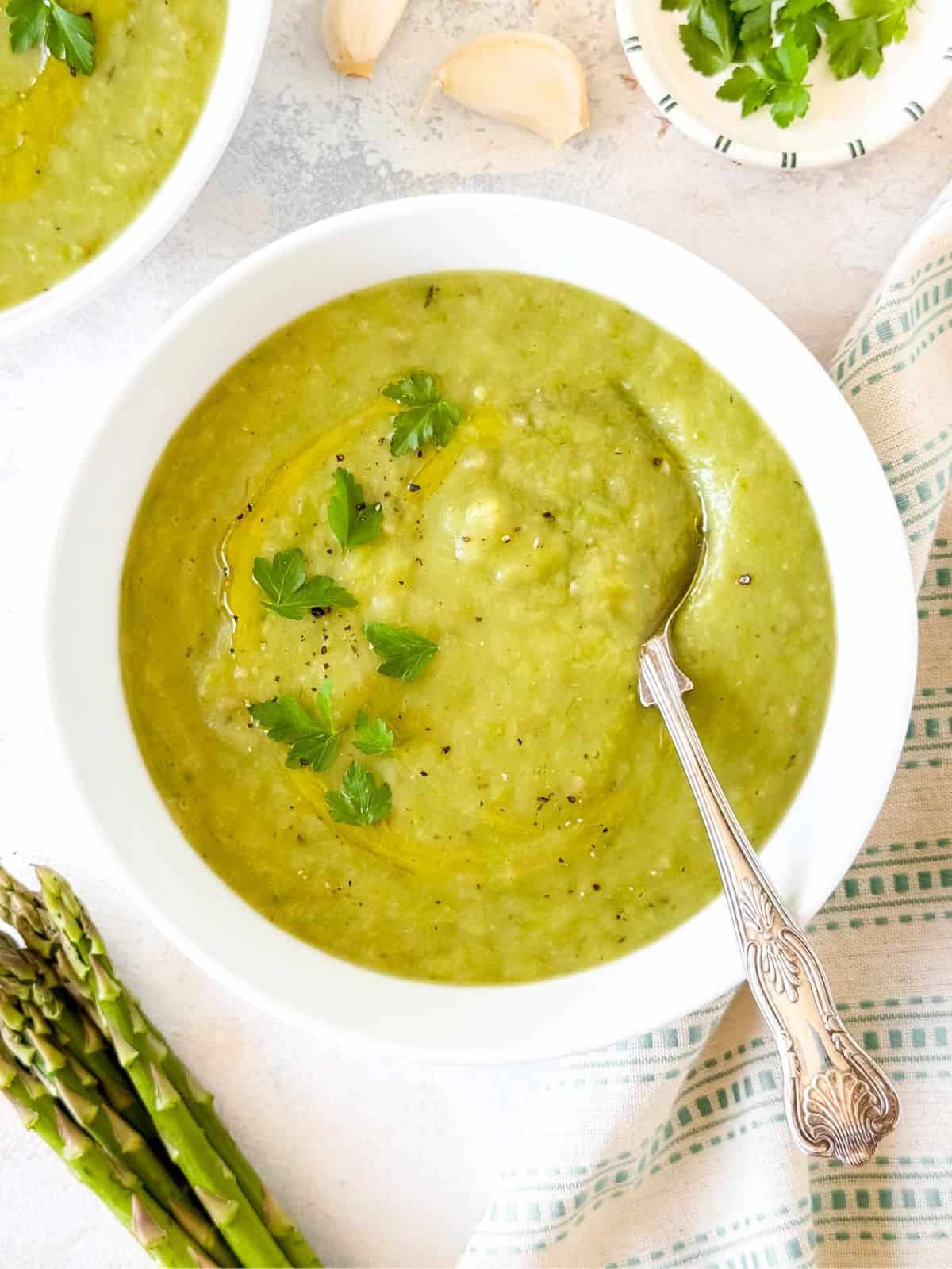 asparagus potato soup in a white bowl next to a bunch of asparagus, garlic and fresh herbs.