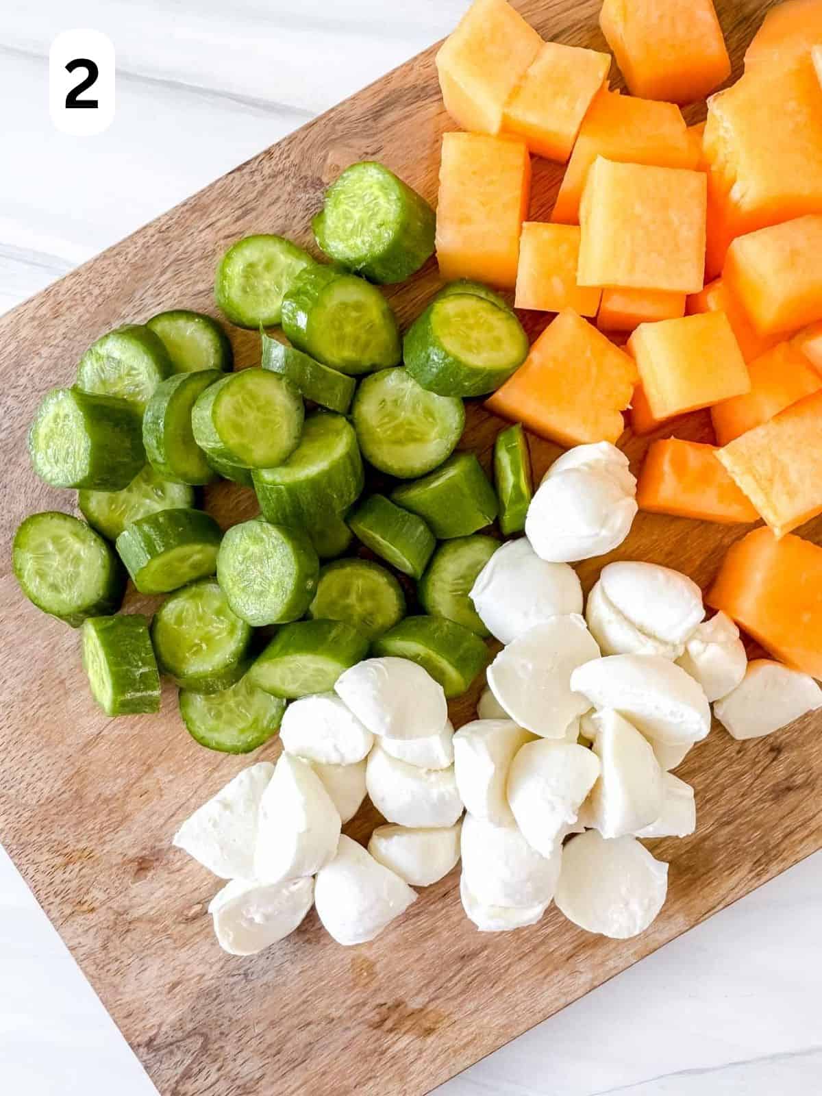diced cucumber, mozzarella and cantaloupe melon on a wooden chopping board labelled number two.
