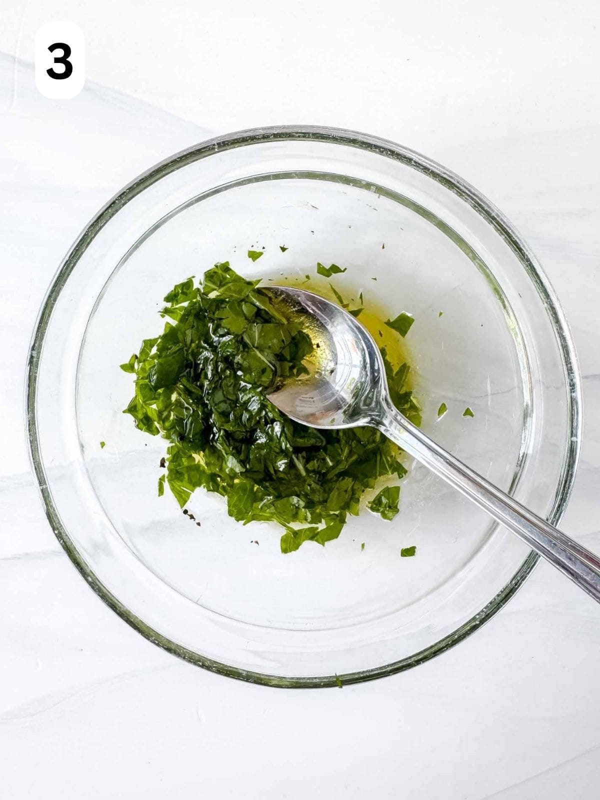 olive oil and diced fresh mint leaves and parsley in a glass bowl with a spoon in it labelled number three.