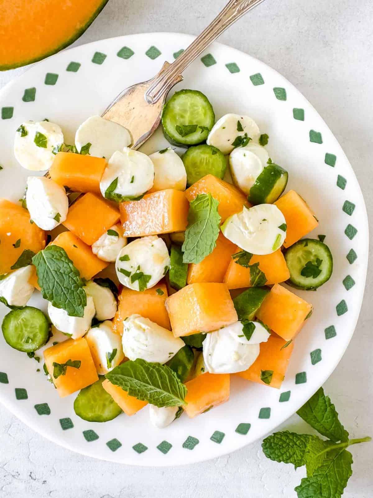 cantaloupe salad with mozzarella and cucumber on a white plate next to fresh mint.