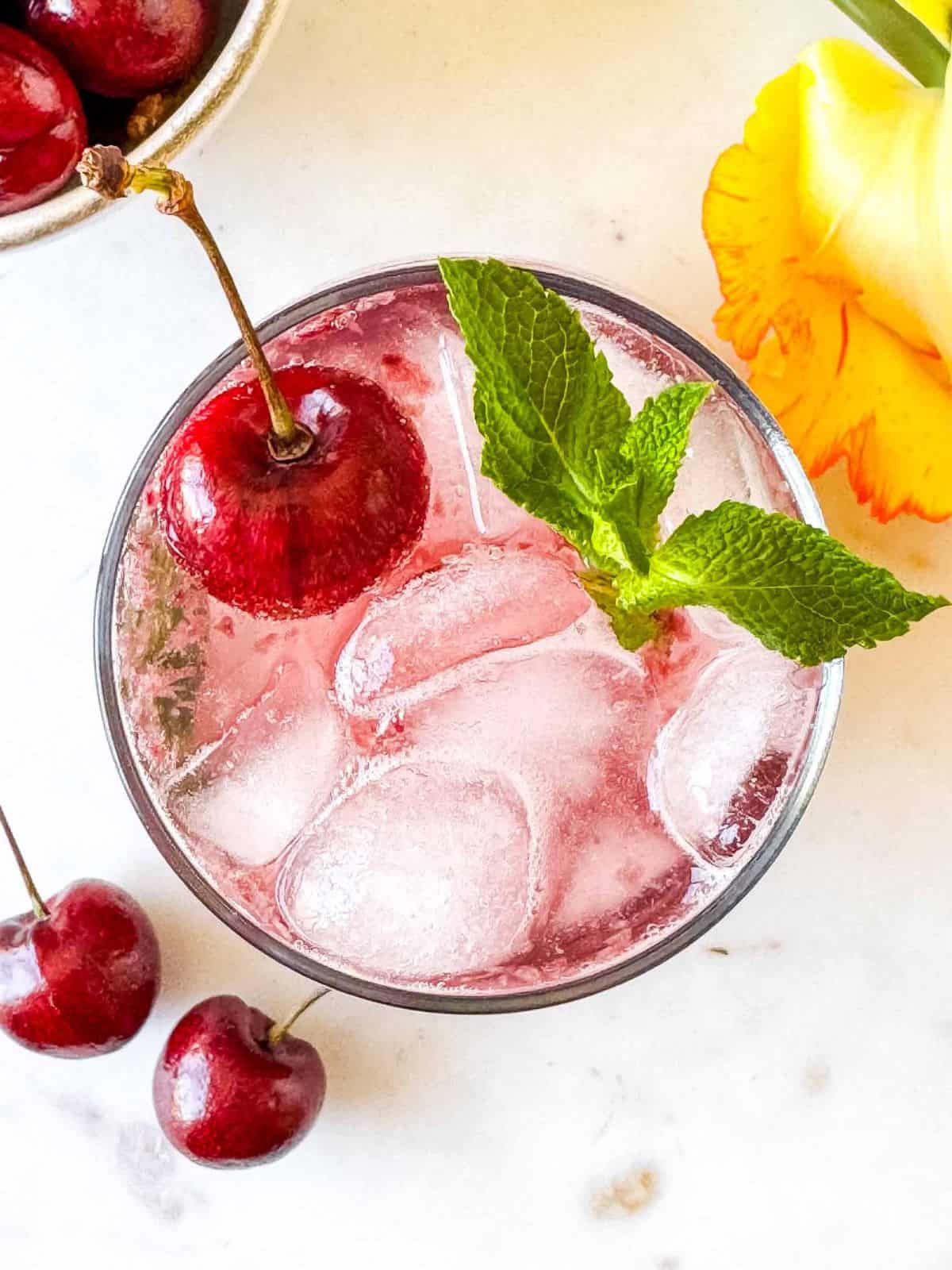cherry mocktail in a glass next to a bowl of cherries and a yellow flower.